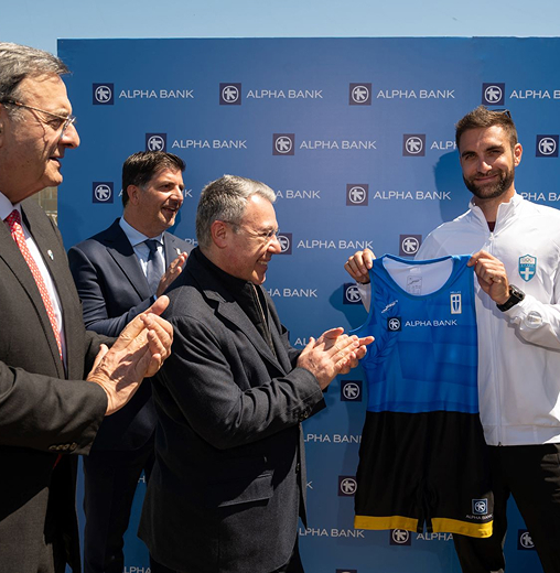 Οι Ισίδωρος Κούβελος, Βασίλης Πολύμερος, Βασίλης Ψάλτης χειροκροτούν αθλητή που κρατά στολή κωπηλασίας με λογότυπο Alpha Bank.