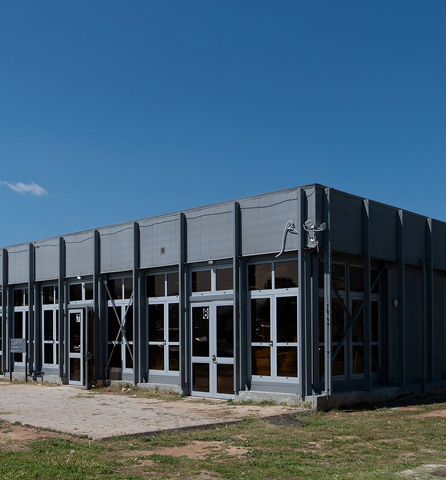 Εξωτερική όψη γκρι, μεγάλου βιομηχανικού κτιρίου με μεταλλική πρόσοψη δίπλα σε υδάτινη έκταση.