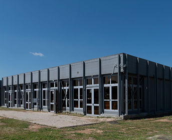 Εξωτερική όψη γκρι, μεγάλου βιομηχανικού κτιρίου με μεταλλική πρόσοψη δίπλα σε υδάτινη έκταση.